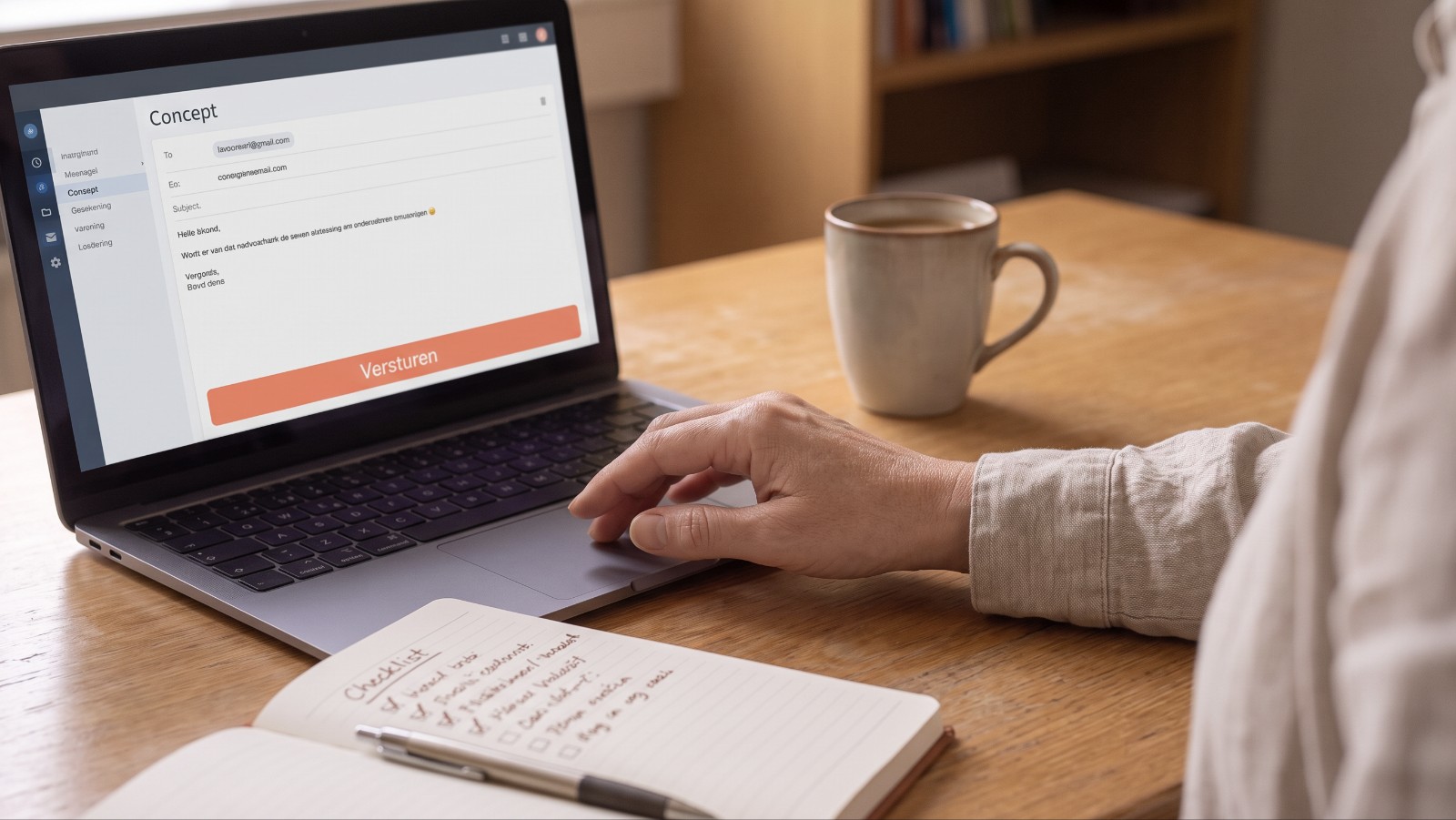 Een hand boven de verzendknop van een conceptmail op een laptop, naast een kop koffie en een notitieblok